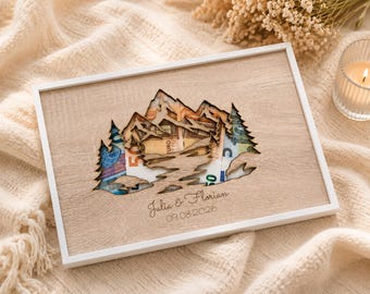 Könnte beinhalten: Ein rechteckiger Holzrahmen mit einem lasergeschnittenen Berg- und Tannenmotiv. Hinter dem Ausschnitt sind Euro-Banknoten sichtbar. Die Holzoberfläche ist mit den Namen Julia & Florian und dem Datum 09.08.2026 in Schreibschrift graviert.