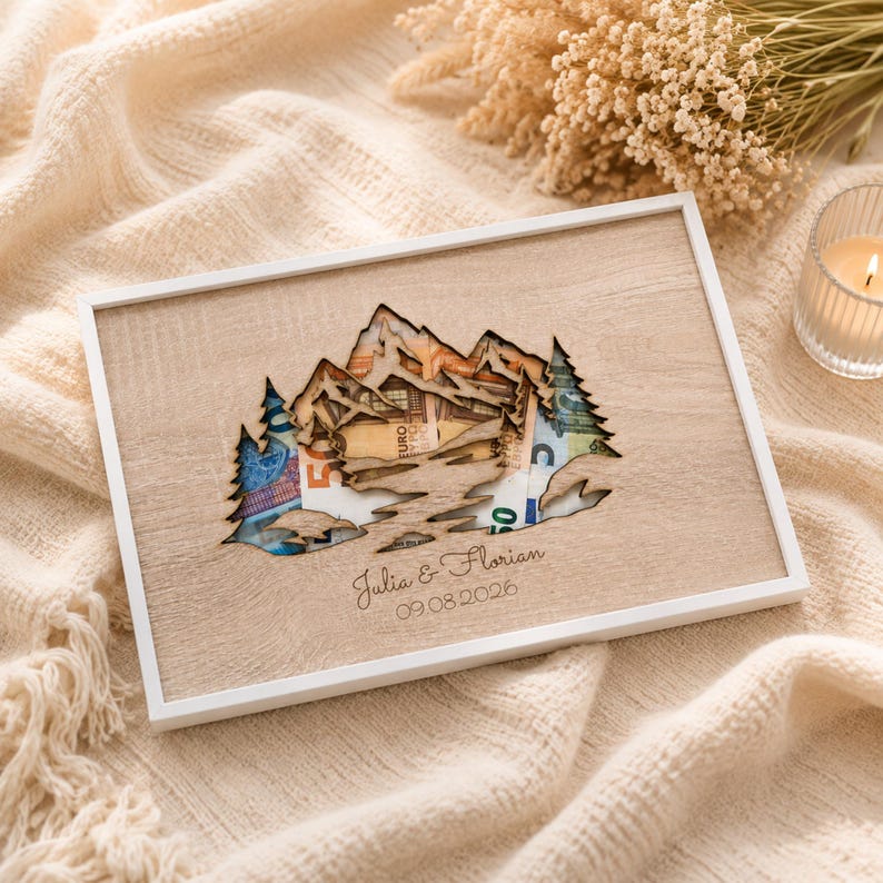 May include: A rectangular wooden frame featuring a laser-cut mountain and pine tree design. Behind the cutout, Euro banknotes are visible. The wood surface is engraved with the names Julia & Florian and the date 09.08.2026 in a script font.