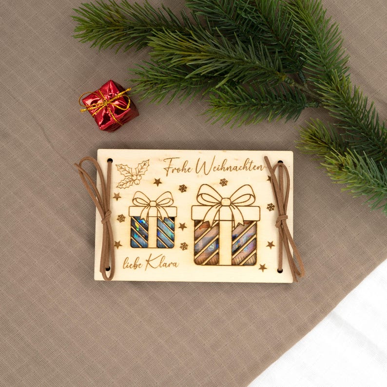 Pu&ograve; includere: Ornamento natalizio in legno con testo inciso al laser e disegni di scatole regalo. L'ornamento ha lacci in pelle marrone ed &egrave; decorato con stelle e agrifoglio. Una piccola confezione regalo rossa e rami di pino sono sullo sfondo.