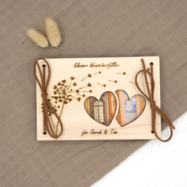 May include: A wooden box with a cutout of two hearts, a dandelion design, and the text "Kleiner Wunscherf&uuml;ller f&uuml;r Sarah & Tom". The box is tied with brown string.
