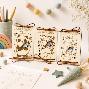 Könnte beinhalten: Drei rechteckige Holzschilder in hellem Farbton mit lasergravierten Tierillustrationen: ein Panda, eine Katze und ein Bär. Jedes Schild zeigt einen Namen, Sterne, Herzen und eine braune Lederschleife. Der Text umfasst Endlich Schulkind, Clara, Du bist 1. Klasse, Lili, Schulkind 2026 und Noah.