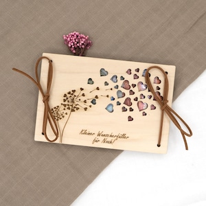 May include: A wooden box with a dandelion design and cut-out hearts. The box has a brown leather cord closure and the text "Kleiner Wunscherf&uuml;ller f&uuml;r Noah!" is engraved on the front.