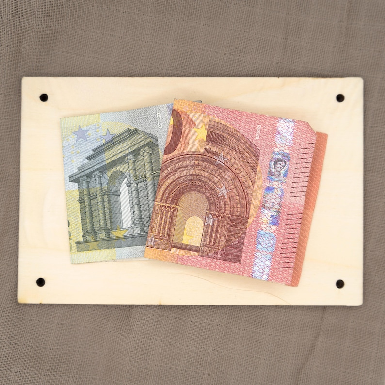 May include: Two Euro banknotes on a light brown wooden surface. The banknotes are both folded in half and show the front side of the bills. The bill on the left is a 20 Euro bill and the bill on the right is a 50 Euro bill.