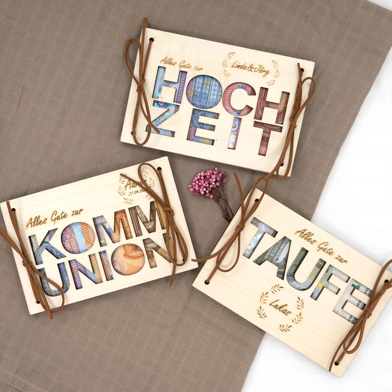 May include: Three wooden boxes with cut-out lettering spelling "Hochzeit", "Kommunion", and "Taufe". Each box has a brown ribbon and is decorated with a laurel wreath. The boxes are designed to hold money as a gift.