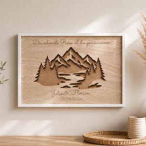May include: A rectangular wooden wall art piece in a white frame featuring a laser-cut layered mountain and forest landscape. The text "Die sch&ouml;nste Reise ist die gemeinsame", "Julia & Florian", and "09.08.2026" is engraved on the light wood surface.
