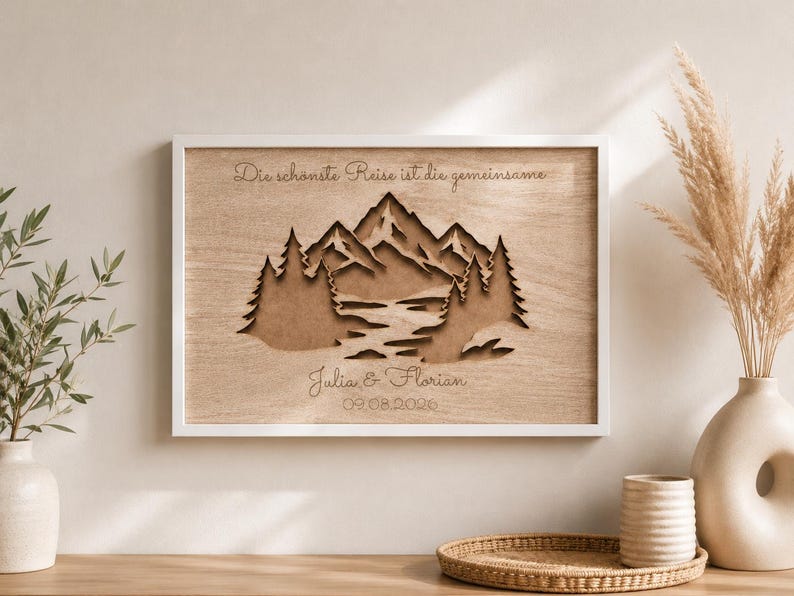 May include: A rectangular wooden wall art piece in a white frame featuring a laser-cut layered mountain and forest landscape. The text "Die sch&ouml;nste Reise ist die gemeinsame", "Julia & Florian", and "09.08.2026" is engraved on the light wood surface.