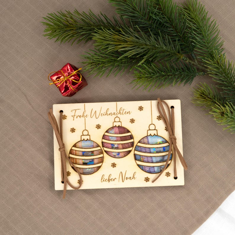 Pu&ograve; includere: Ornamento natalizio in legno con tre palline colorate, lacci in pelle marrone e la scritta "Frohe Weihnachten lieber Noah". Un piccolo cofanetto rosso e rami di pino sullo sfondo.