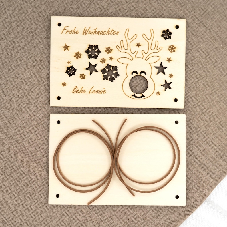 Pu&ograve; includere: Due decorazioni natalizie in legno. L'ornamento superiore presenta un disegno di renna con le parole "Frohe Weihnachten" e "liebe Leonie" incise nel legno. L'ornamento inferiore ha due corde marroni.