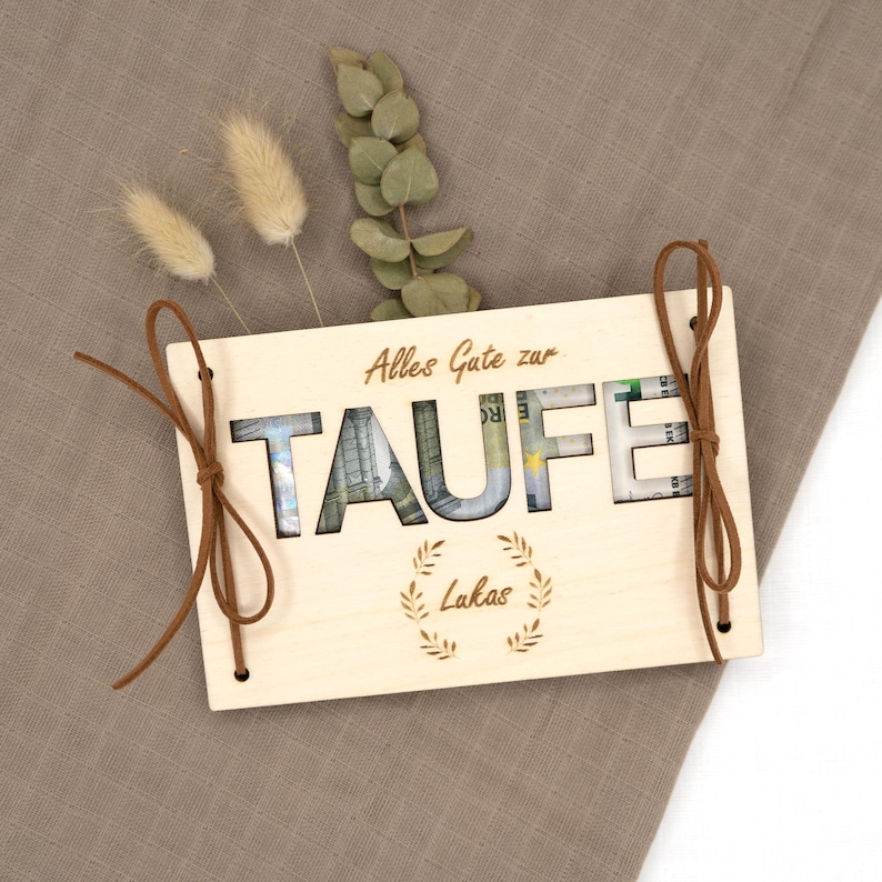 Puede incluir: Una caja de madera con un recorte de la palabra "TAUFE" con dinero insertado en los recortes. La caja est&aacute; atada con una cinta marr&oacute;n y tiene el texto "Alles Gute zur Taufe" y "Lukas" en ella.