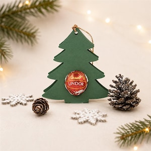 Può includere: Un addobbo in carta verde scuro a forma di albero con un ritaglio circolare al centro che contiene un singolo tartufo al cioccolato Lindt Lindor rosso e sferico. L'ornamento include un sottile filo dorato nella parte superiore per appenderlo.
