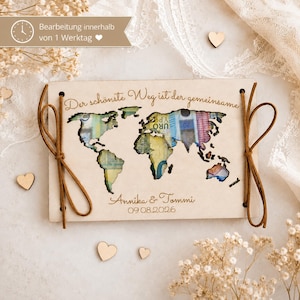 Può includere: Una targa rettangolare in legno chiaro con una sagoma del mondo tagliata al laser. L'intaglio rivela banconote colorate sottostanti. Il testo recita "Der schönste Weg ist der gemeinsame", "Annika & Tommi" e "09.08.2026". Cordini in pelle marrone sono annodati attraverso i fori sui lati sinistro e destro.