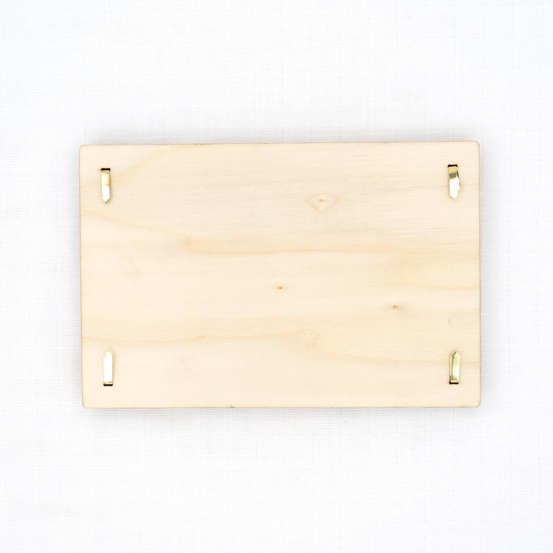 Pu&ograve; includere: Una targa rettangolare in legno di colore chiaro con una finitura a venature naturali e liscia. La targa presenta quattro piccole clip metalliche color oro posizionate vicino a ogni bordo, progettate per fissare oggetti sulla superficie.