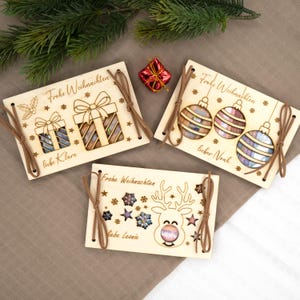Pu&ograve; includere: Tre biglietti di Natale in legno con disegni tagliati al laser e lacci di spago marrone. Le carte presentano il testo "Frohe Weihnachten" e nomi personalizzati. Un biglietto ha disegni di scatole regalo, un altro ornamenti e l'ultimo una renna.