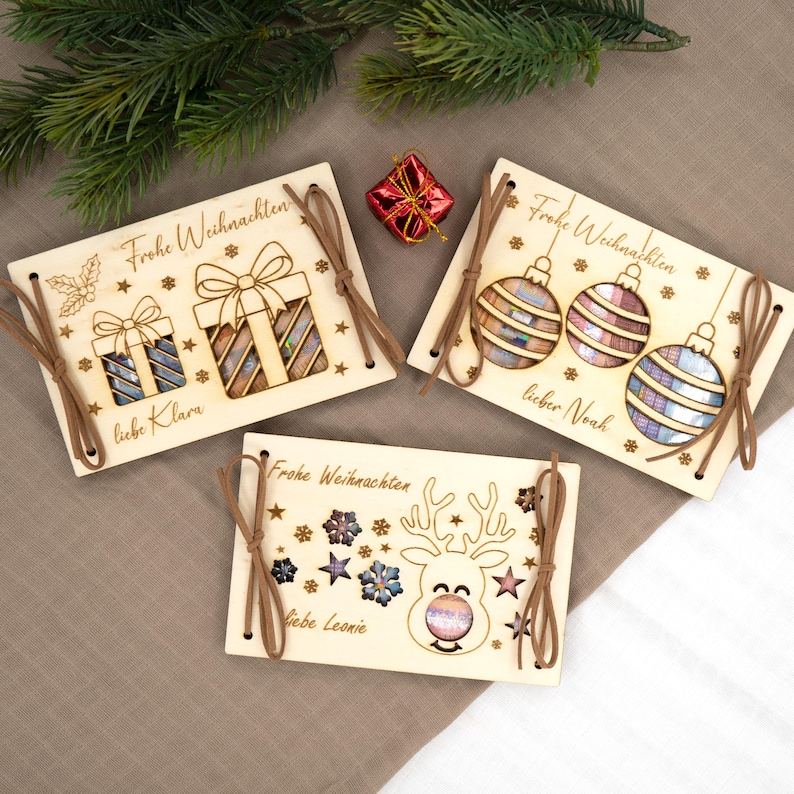 Pu&ograve; includere: Tre biglietti di Natale in legno con disegni tagliati al laser e lacci di spago marrone. Le carte presentano il testo "Frohe Weihnachten" e nomi personalizzati. Un biglietto ha disegni di scatole regalo, un altro ornamenti e l'ultimo una renna.
