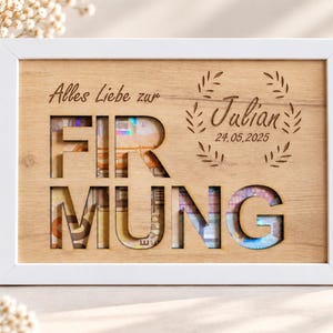 May include: A rectangular wooden frame with a white border containing a light wood panel. The panel features the laser-cut text "FIRMUNG" with visible currency behind the cutouts. It includes the engraved text "Alles Liebe zur Julian 24.05.2025" and a leaf wreath design.