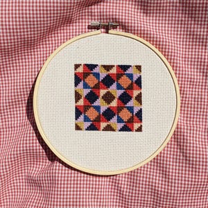 Pode incluir: Um projeto de ponto cruz concluído num bastidor de bordar de madeira. O design apresenta um padrão geométrico de diamantes coloridos em tons de vermelho, azul, laranja e castanho. O bastidor está sobre um tecido xadrez vermelho e branco.