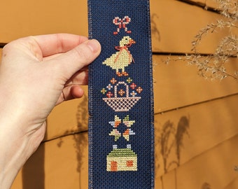 May include: A blue fabric bookmark featuring a stitched design of a yellow duck with a red bow, a basket of flowers, a star, and a small house.