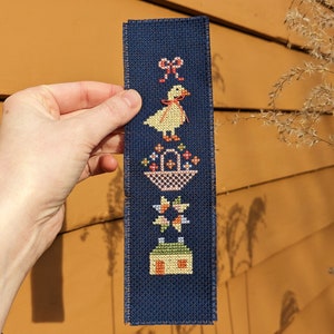 Pode incluir: Um marcador de página de tecido azul com um desenho bordado de um pato amarelo com um laço vermelho, uma cesta de flores, uma estrela e uma pequena casa.