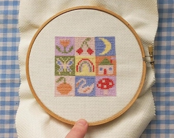 May include: A cross-stitch embroidery hoop with a colorful patchwork design featuring a butterfly, a rainbow, a house, a mushroom, a crescent moon, and a flower. The hoop is made of wood and has a white fabric background.