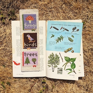 Könnte beinhalten: Nahaufnahme eines Buches, das auf einer Seite über die Birkenfamilie aufgeschlagen ist. Ein farbenfrohes Lesezeichen mit einem Kreuzstich-Design aus einem Pilz, einem Vogel und einem Baum ragt aus dem Buch heraus. Das Lesezeichen trägt die Wörter "fungi", "birds" und "trees".