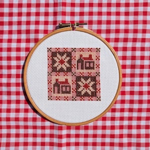 May include: A circular wooden embroidery hoop containing a piece of white Aida fabric with a cross-stitch pattern. The design features four alternating squares in shades of brown, pink, and red, depicting two stylized houses and two star motifs.