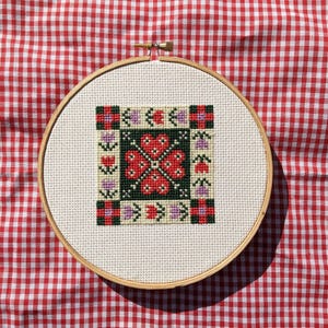 May include: A red, green and purple cross-stitch design in a wooden embroidery hoop. The design features a floral pattern with a central motif of four red hearts.