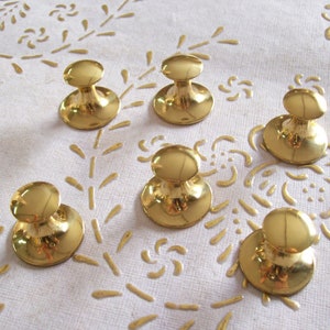 Puede incluir: Seis pomos de metal dorado con parte superior redonda y base redonda. Los pomos están dispuestos en semicírculo sobre una superficie blanca.