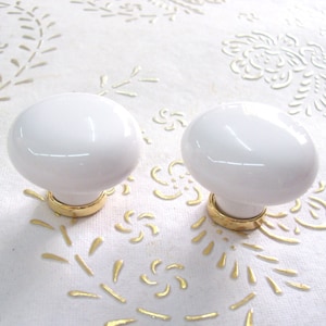 Może przedstawiać: Dwa białe ceramiczne gałki do szafek z podstawami z metalu w kolorze złotym.