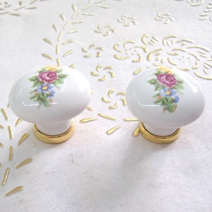 May include: Two white ceramic cabinet knobs with gold trim. Each knob features a floral design with pink, yellow, and blue flowers.