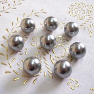 Può includere: Un gruppo di nove bottoni color argento a forma di perla su una superficie bianca con un motivo floreale dorato.