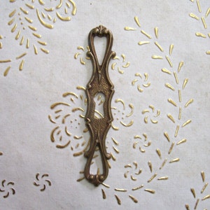 Può includere: Un copri-chiave in ottone con un design decorativo e ornato. Il copri-chiave è di colore oro chiaro e ha un buco della serratura al centro.