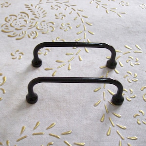 Peut inclure: Deux poignées d'armoire noires et arquées avec une finition mate. Elles sont posées sur une surface blanche avec un motif floral doré. Les poignées sont conçues pour une installation facile et ajoutent de l'élégance.
