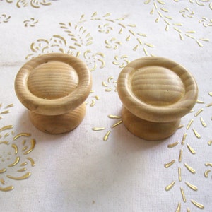 以下が含まれることがあります： 未完成の木製キャビネットノブ2つ。 各ノブは、丸く溝のある上部と円筒形のベースを備えています。 ノブは薄茶色で、金色の花のアクセントが付いた白い表面にあります。 家具の修復プロジェクトに最適です。