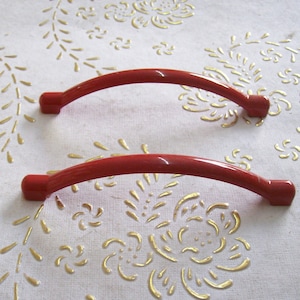 May include: Two red plastic cabinet door handles. The handles are curved and have a smooth finish.