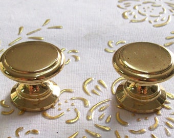 May include: Two gold-toned cabinet knobs with a round, slightly raised top and a smaller, round base. The knobs have a smooth, polished finish.