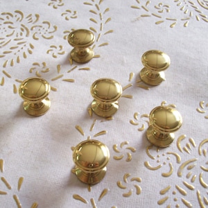 May include: Six gold-toned cabinet knobs with a round shape and a raised centre. The knobs are arranged in a semi-circle on a white surface with a floral pattern.
