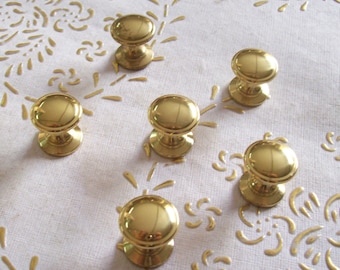 Puede incluir: Seis pomos de armario de color dorado con forma redonda y un centro elevado. Los pomos están dispuestos en semicírculo sobre una superficie blanca con un patrón floral.