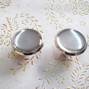 May include: Two silver round cabinet knobs with a raised edge. The knobs are on a white background with a gold floral pattern.