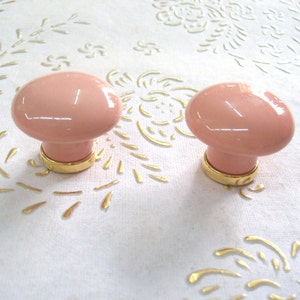Op de afbeelding: Twee roze keramische kastknoppen met gouden metalen bases.
