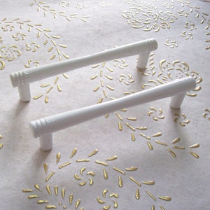 May include: Two white metal cabinet drawer pulls with a simple, rectangular design. The pulls are on a white surface with a gold floral pattern.