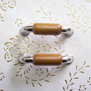 May include: Two chrome and wood cabinet pulls with a brown wood finish. The pulls are on a white background with a gold floral pattern.