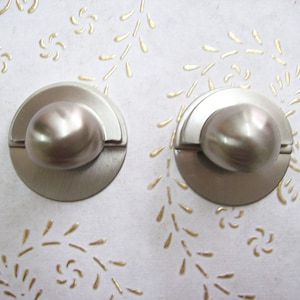 Peut inclure: Deux boutons de cabinet argentés avec un centre rond légèrement surélevé et un design en demi-cercle sur le côté.