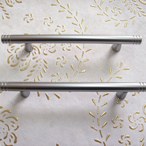 May include: Two brushed nickel cabinet pulls with a cylindrical design and grooved ends. The pulls are mounted on a light-coloured surface with a gold floral pattern. Suitable for kitchen or bathroom cabinets.