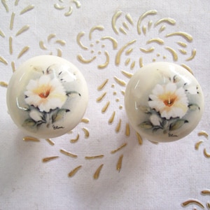 Può includere: Due pomelli in ceramica bianca con un design floreale dipinto. Ogni pomello presenta un fiore bianco con un centro giallo e foglie verdi. Lo sfondo dei pomelli è grigio chiaro.