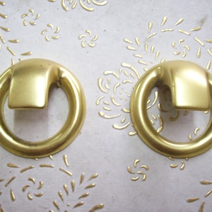 Op de afbeelding: Twee gouden metalen ringgrepen met een rechthoekige vorm in het midden. De ringgrepen bevinden zich op een witte achtergrond.