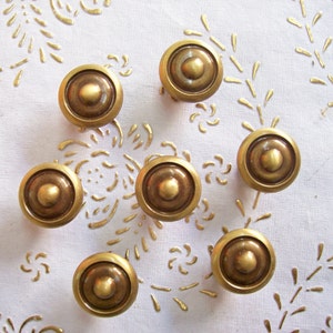 Peut inclure: Huit boutons de porte en métal doré avec un centre surélevé. Les boutons sont disposés en demi-cercle sur une surface blanche.