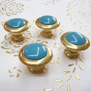 May include: Four gold cabinet knobs with a turquoise blue centre. The knobs are round and have a raised edge around the centre.