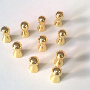 Puede incluir: Diez pomos de metal de color dorado con una parte superior redonda y una base ligeramente cónica. Los pomos están dispuestos en un patrón suelto sobre una superficie blanca.