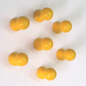 Puede incluir: Siete chinchetas de plástico amarillo con cabeza redonda. Las chinchetas están dispuestas de forma dispersa sobre una superficie blanca.