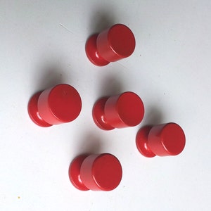 Peut inclure: Cinq boutons en plastique rouge avec un dessus rond et une petite base. Les boutons sont disposés en forme d'étoile sur une surface blanche.